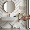 Gold-framed round mirror above white marble bianco dolomite chevron mosaic vanity with gold geometric accents