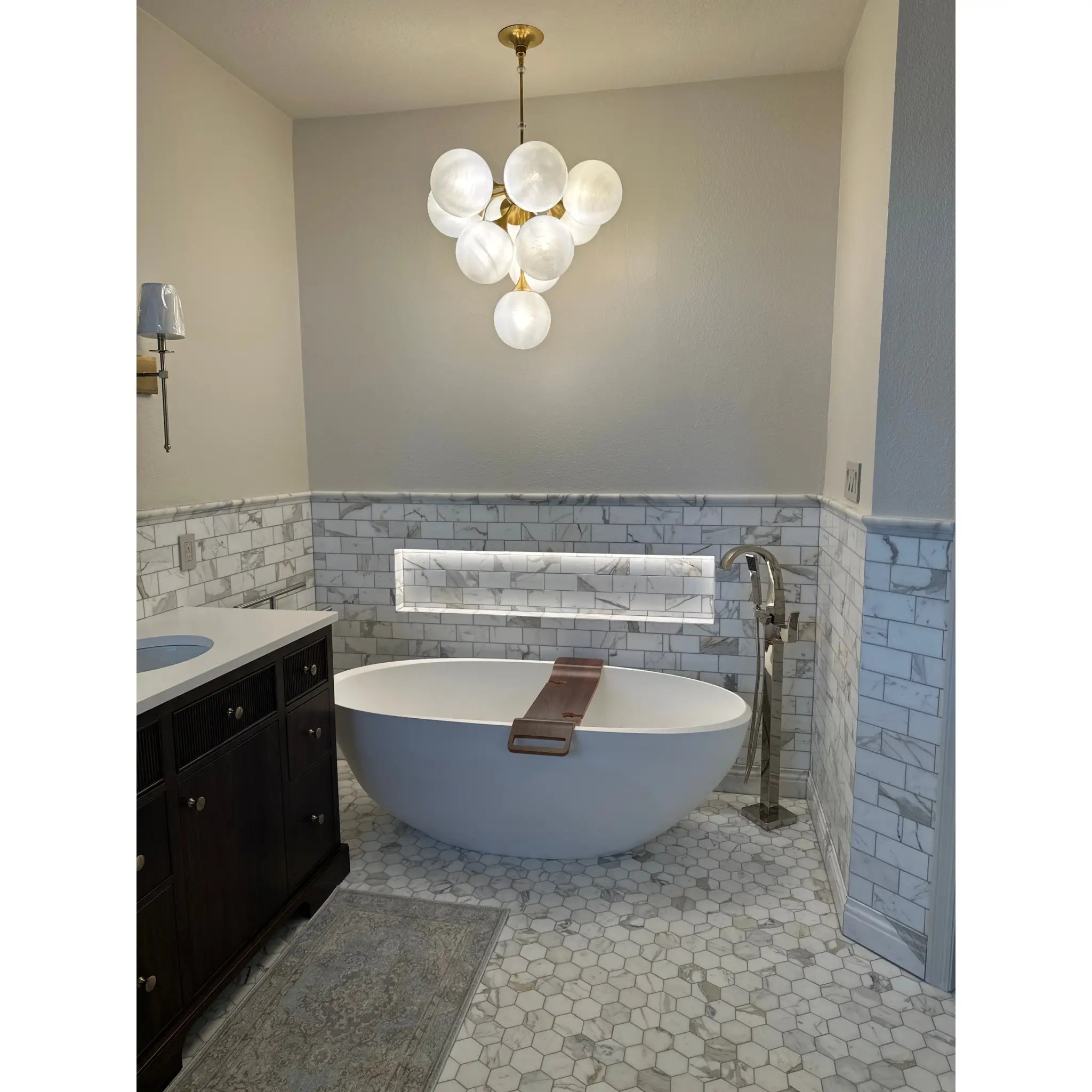 White freestanding bathtub with a brown wooden bath mat shown alongside calacatta gold 2 inch hexagon mosaic marble tile polished or honed