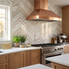 Copper range hood with sleek angular design and brushed metal finish paired with calacatta gold chevron mosaic tile