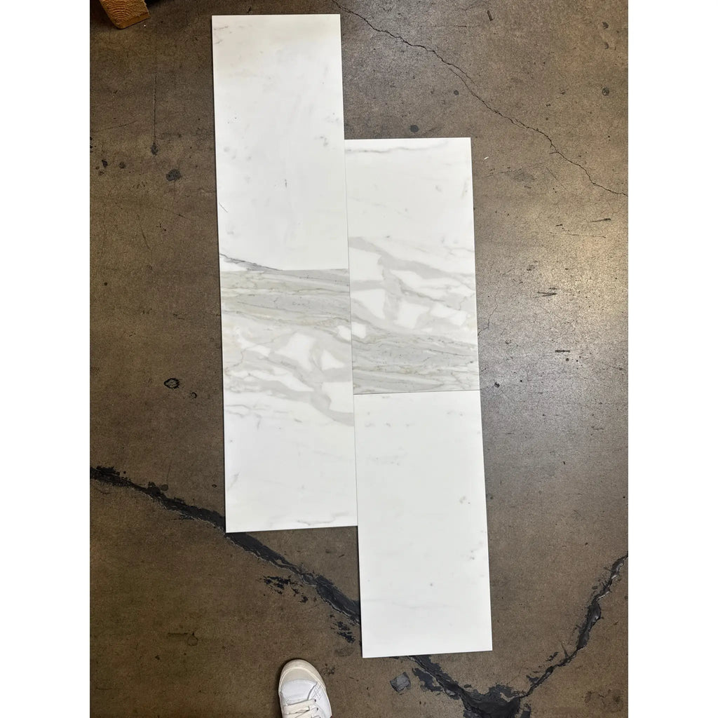 White and gray marble-look tile samples on dark floor for Calacatta Gold Marble 12X24 product