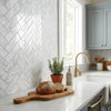 A rustic wooden cutting board with natural grain holding a round loaf and slices beside carrara white herringbone marble tile