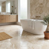 Free-standing white bathtub with gold-toned fixtures and towel over side on Diano Royal queen beige marble hexagon mosaic tile