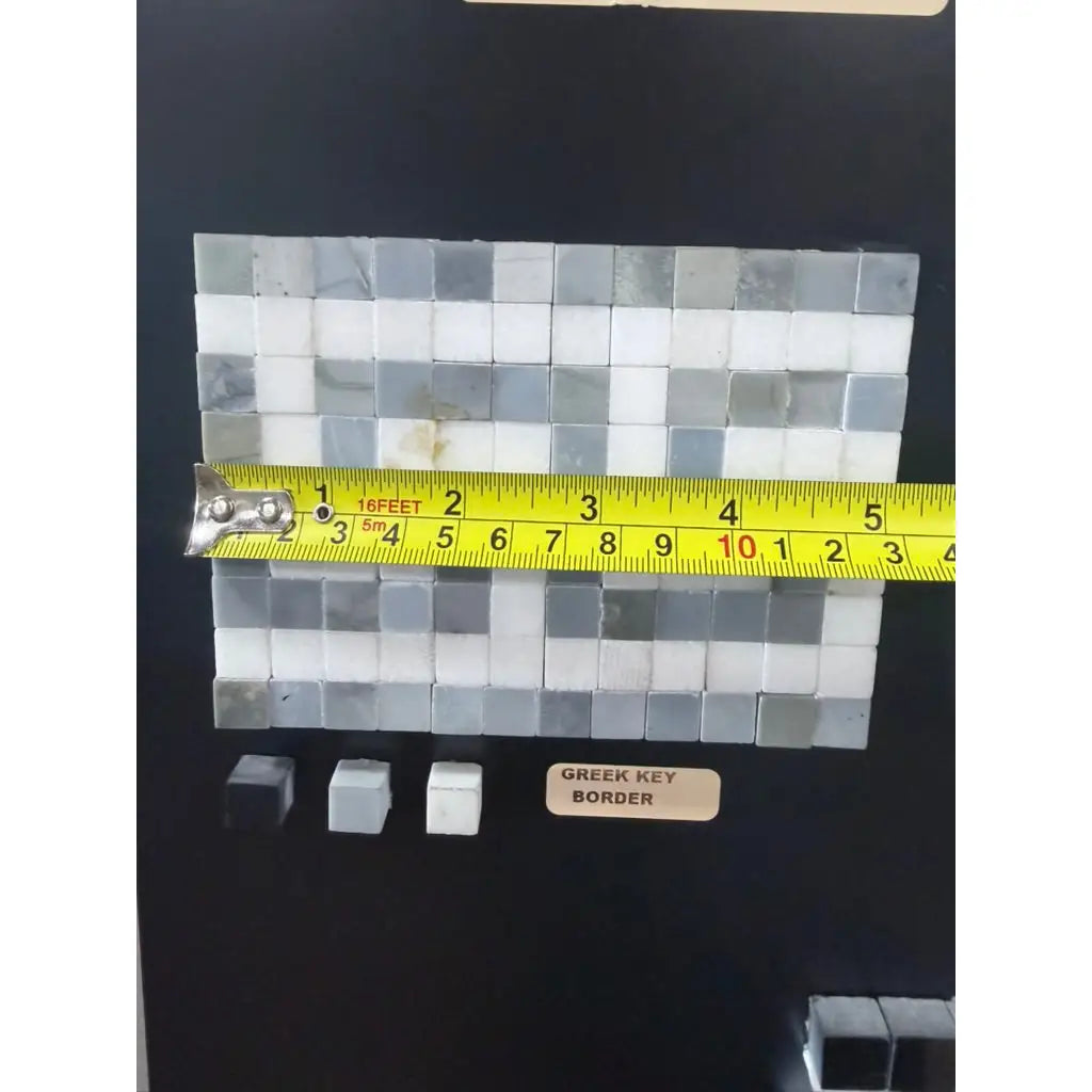 A rectangular thassos white marble mosaic tile panel with greek key border in shades of white and gray with measuring tape