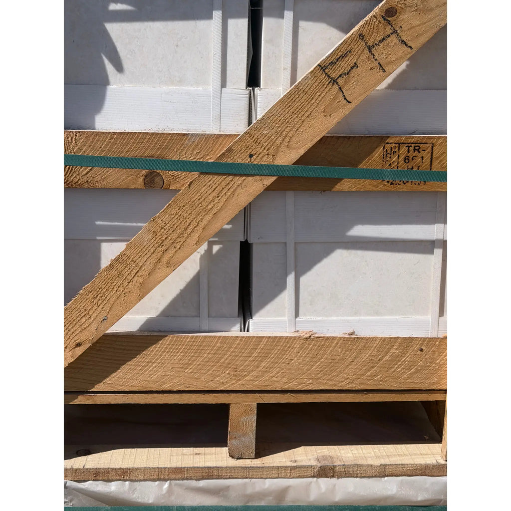 A light brown wooden pallet with a green strap securing white pearl botticino beige marble 12x12 polished or honed tiles