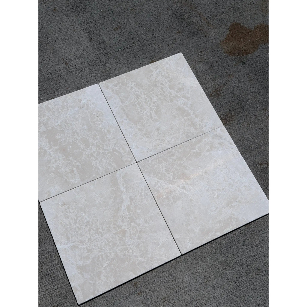 Four light gray white pearl botticino beige marble 12x12 tumbled tiles with subtle veining on dark concrete surface